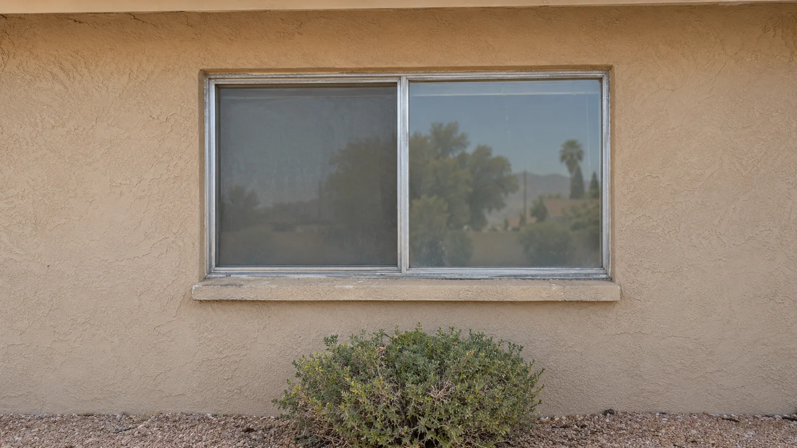 Old oxidized aluminum sliding window with hazy glass in tan stucco, a shrub below