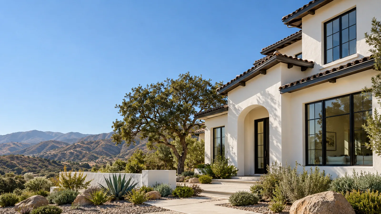White stucco Spanish-style home with black-framed windows and an arched doorway, an oak tree and a succulent yard