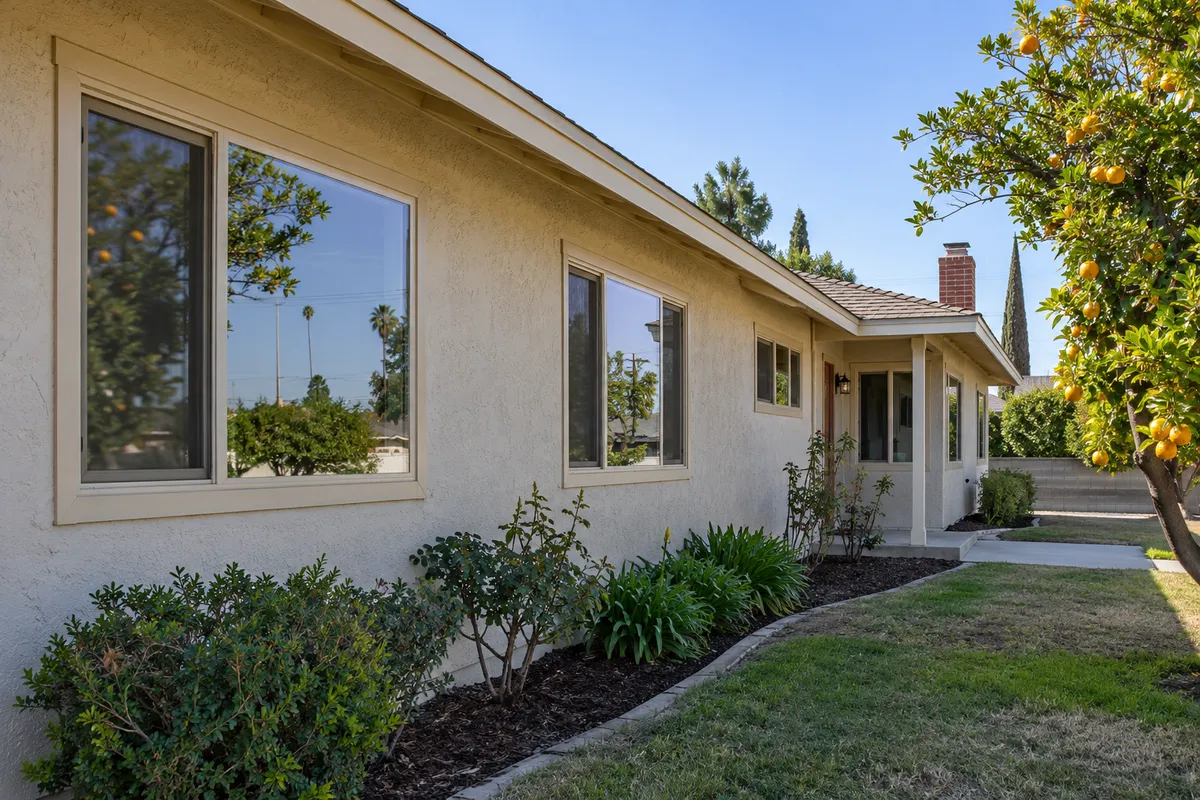 Long single-story stucco ranch wall with large new beige-framed retrofit windows reflecting an orange tree